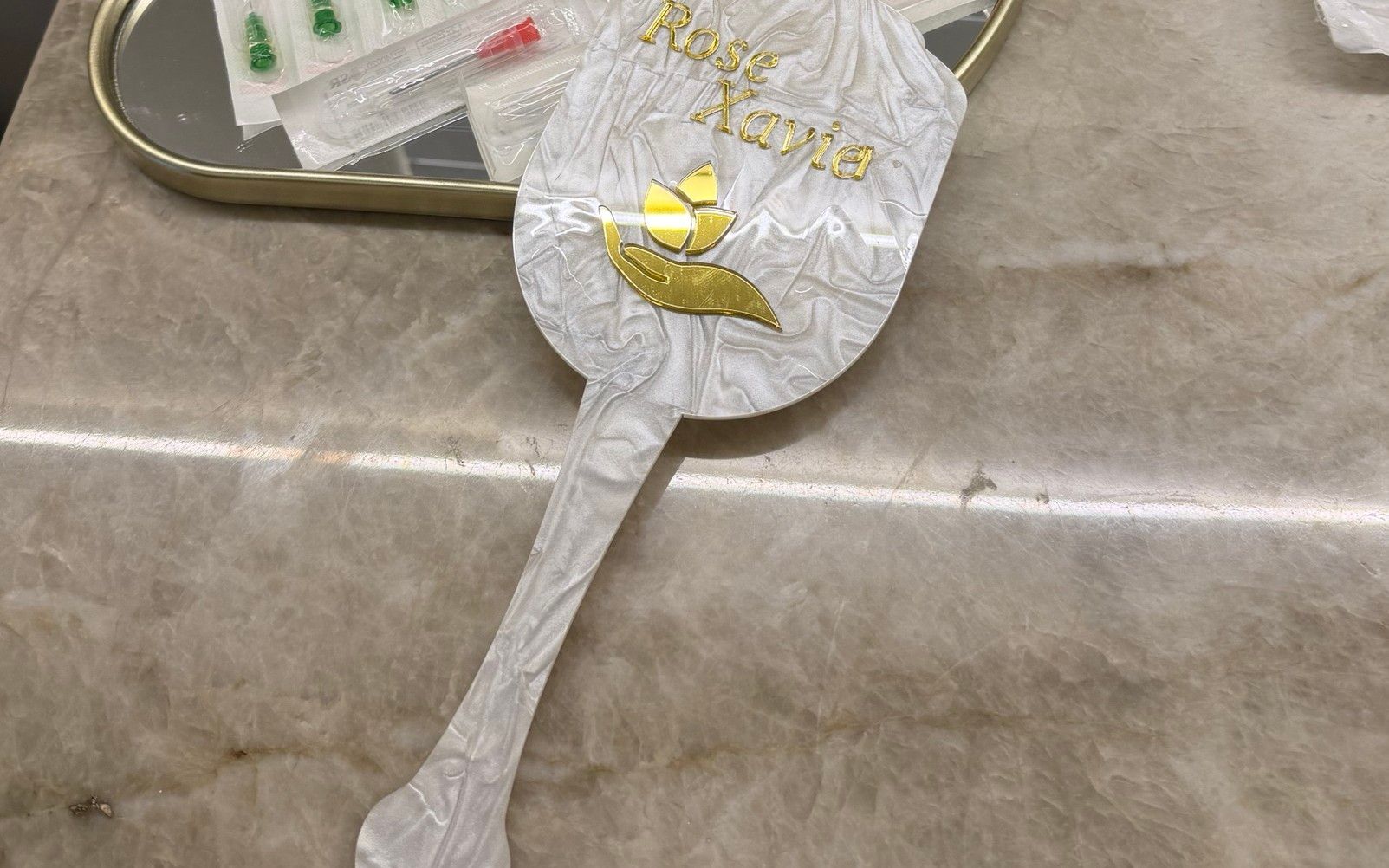 Bandeja espelhada com seringas lacradas e o espelho de mão perolado com o nome Rose Xavier em dourado, sobre a bancada de pedra do consultório.