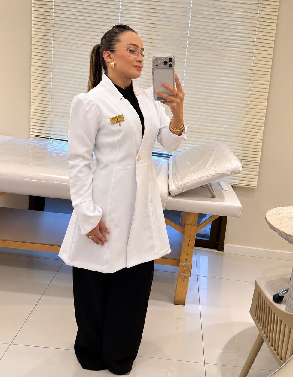 Rose Xavier de jaleco branco acinturado, fazendo uma foto no espelho do consultório.
