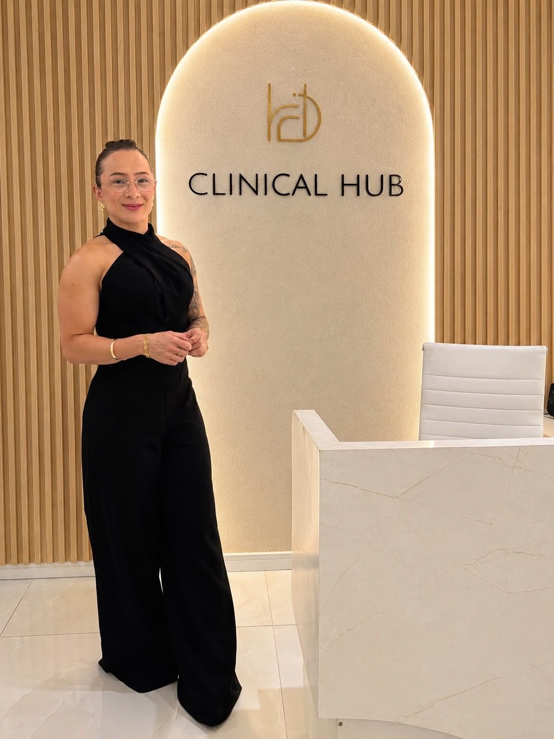 Rose Xavier, de macacão preto, em pé na recepção do Clinical Hub, ao lado do arco iluminado com o nome da clínica.